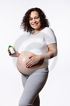 pregnant woman smiles at camera, applying stretch marks cosmetic oil, massaging her big belly, white isolated background