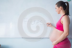 A pregnant woman in sportswear writes the word fitness on a white marker board. A girl in the 3rd trimester goes in for