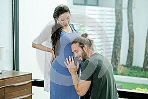 Pregnant woman stand with her husband who sit and listen their baby sound in womb. They are happy together to stay at home in