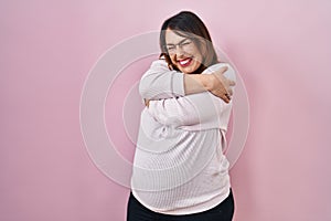 Pregnant woman standing over pink background hugging oneself happy and positive, smiling confident