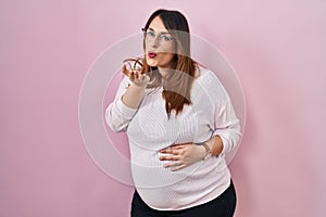 Pregnant woman standing over pink background looking at the camera blowing a kiss with hand on air being lovely and sexy