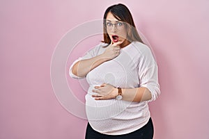 Pregnant woman standing over pink background looking fascinated with disbelief, surprise and amazed expression with hands on chin