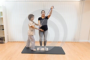 A pregnant woman standing on a proprioceptive trunk or wooden roller with an elastic band with physiotherapist