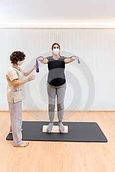 A pregnant woman standing on a proprioceptive trunk or wooden roller excersing with an elastic band with physiotherapist