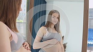 A pregnant woman stands in front of a mirror and holds her hands on her belly. Skin care for pregnant women. Prevention