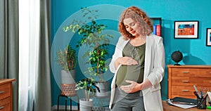 Pregnant woman stands in middle of office room enjoying moment to herself, stroking her belly,
