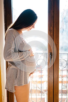 Pregnant woman stands near the balcony door stroking her belly