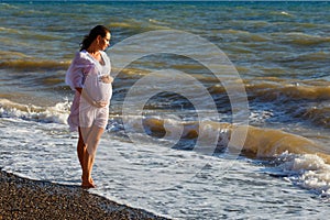 Pregnant woman stands on sea beach in summer in white light dress with fluffy