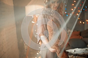 Pregnant woman stands with teddy bear in open peignoir. View through transparent curtain.