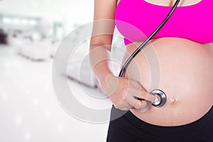 pregnant woman with stethoscope listening belly to baby in hospital background