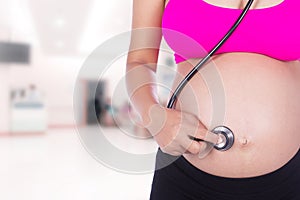 pregnant woman with stethoscope listening belly to baby in hospital background