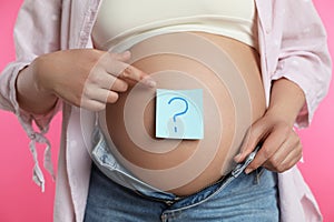 Pregnant woman with sticky note on belly against pink background, closeup. Choosing baby name