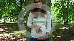 Pregnant woman stroking caress her belly standing near big tree trunk in park