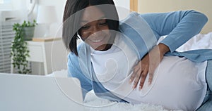 Pregnant woman stroking her belly and using laptop on bed