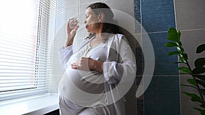 Pregnant woman stroking her big belly, drinking water standing by window in the bathroom.