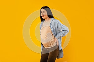 Pregnant woman suffering from pain, having spasm during painful contractions, touching back over yellow background