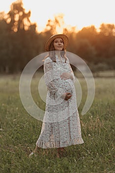 A pregnant woman in a sunset background is holding her belly near the farm