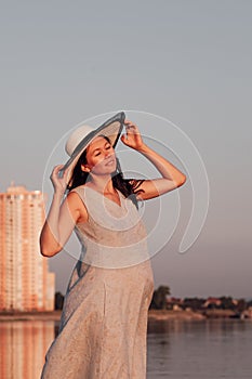 A pregnant woman at sunset. A peaceful, relaxed pregnant woman holds the brim of a straw hat with her hands and enjoys