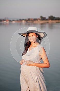 A pregnant woman at sunset. A peaceful, relaxed pregnant woman in a straw hat and dress with her hands on her pregnant