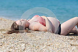 Pregnant woman in swimsuit sunbathing on pebble beach in early time