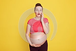 Pregnant woman talking by mobile phone while standing isolated over yellow background, wearing red t shirt, touching her bare