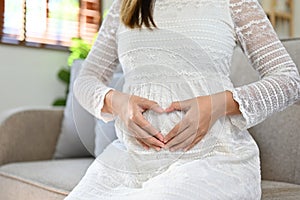 Pregnant woman touching her belly with love, making a heart sign with hands while sitting on sofa