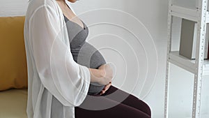 Pregnant woman touching her tummy by hand gently while sitting at sofa near window at home bedroom. People lifestyles and