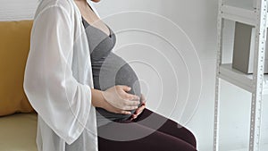 Pregnant woman touching her tummy by hand gently while sitting at sofa near window at home bedroom. People lifestyles and