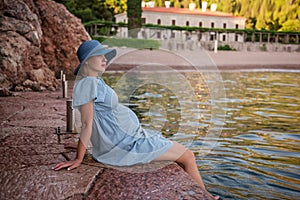 Pregnant woman traveler in denim dress, blue straw hat sits by the sea, dangles legs in water