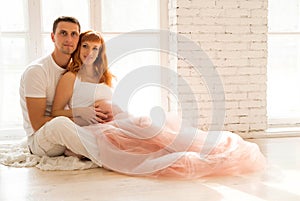 Pregnant woman in a tulle skirt and her husband are sitting on the floor near a large window