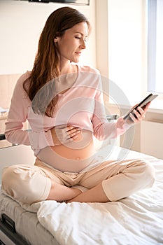 Pregnant woman using cellphone resting on bed in prenatal center