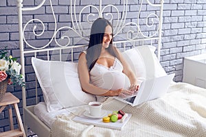Pregnant woman using laptop and having breakfast in bed