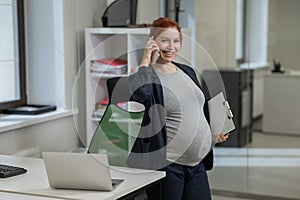 Pregnant woman using mobile phone and holding paper tablet in office.