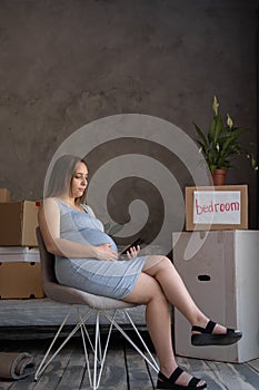 Pregnant woman using tablet sitting at home in armchair after moving house, owner of new apartment resting with digital
