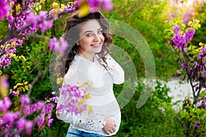 Pregnant woman walking in the autumn park. With one hand she hugs belly and touches flowers with other, she is looking