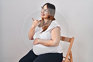 Pregnant woman wearing band aid for vaccine injection pointing thumb up to the side smiling happy with open mouth