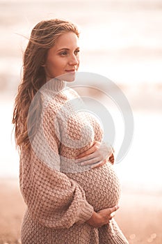 Pregnant woman wearing white dress sittin over sea shore outdoors. Motherhood. Maternity. Healthy lifestyle
