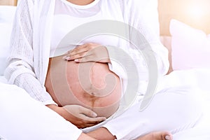 pregnant woman in white dress with hand on her belly in bedroom. Young woman holding hands on the belly of pregnant