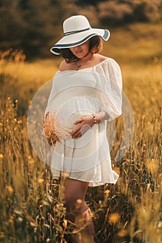 A pregnant woman in a white dress and hat stands elegantly in a golden wheat field, radiating maternal serenity
