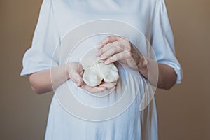 Pregnant woman in white dress holding baby socks with her hands near belly