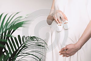 Pregnant woman in white dress holding small shoes for unborn baby on belly, preparing go to maternity hospital for