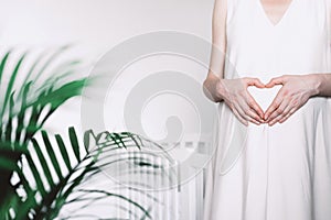 Pregnant woman in white dress making heart on pregnant belly. Maternity, preparation for childbirth, baby
