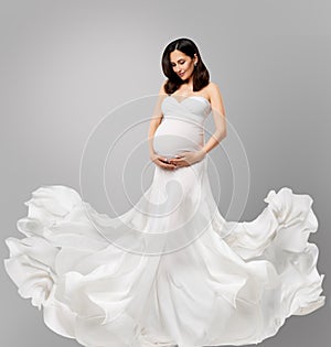 Pregnant Woman in White Long Dress flying on Wind looking at Belly. Beautiful Mother in Silk waving Gown over Gray Background.
