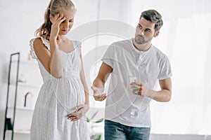 pregnant woman in white nightie having headache while husband giving medicines and glass of water to her