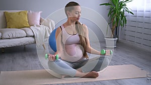 Pregnant woman working out with dumbbells and sitting on floor in apartment room spbd.