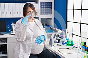 Pregnant woman working at scientist laboratory pointing with hand finger to face and nose, smiling cheerful