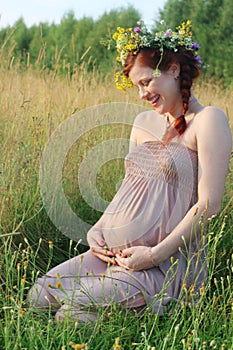 Pregnant woman in wreath sits and laughs in dry field