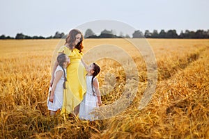 A pregnant woman in a yellow dress with her daughter in white sundresses stand hugging each other in a wheat field looking at each