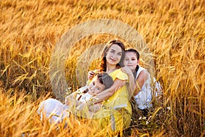 A pregnant woman in a yellow dress with two daughters in white sundresses sit hugging in a wheat field