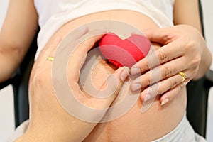 Pregnant women hand and husband hand keep a red heart symbol placed on belly concept of love for the baby.